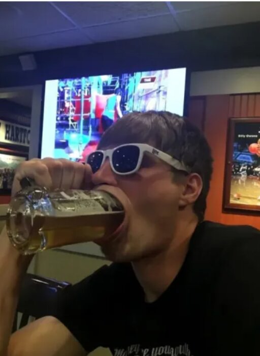 En person med solglasögon dricker öl ur en mugg i en bar. TV-skärmar syns i bakgrunden.