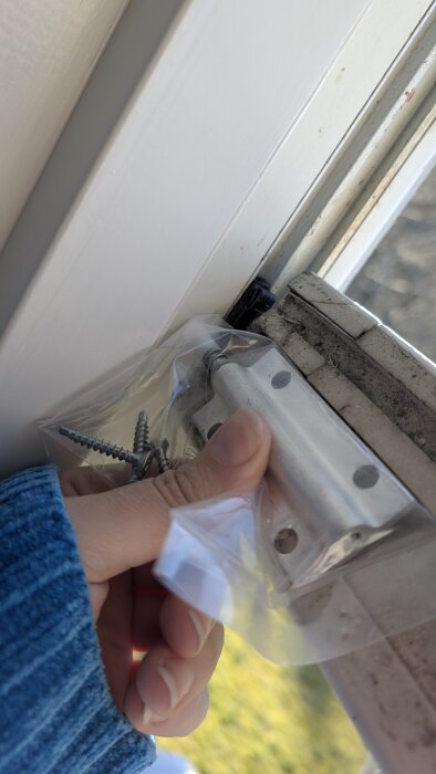 Hand som håller ett paket med ett fönsterbeslag och skruvar nära ett öppet fönster. Beslaget verkar inte passa på grund av höjden.