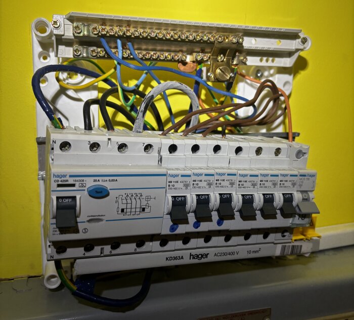 Elcentral med jordfelsbrytare och flera säkringar installerade i en vägg med gul bakgrund, flera elkablar är anslutna.