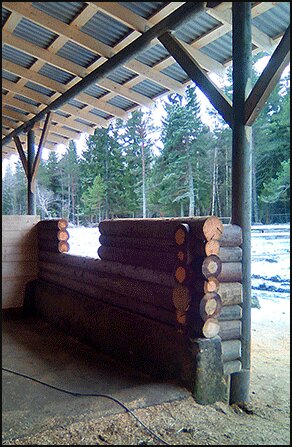 Round log wall construction under a covered outdoor area, with a snowy forest backdrop.