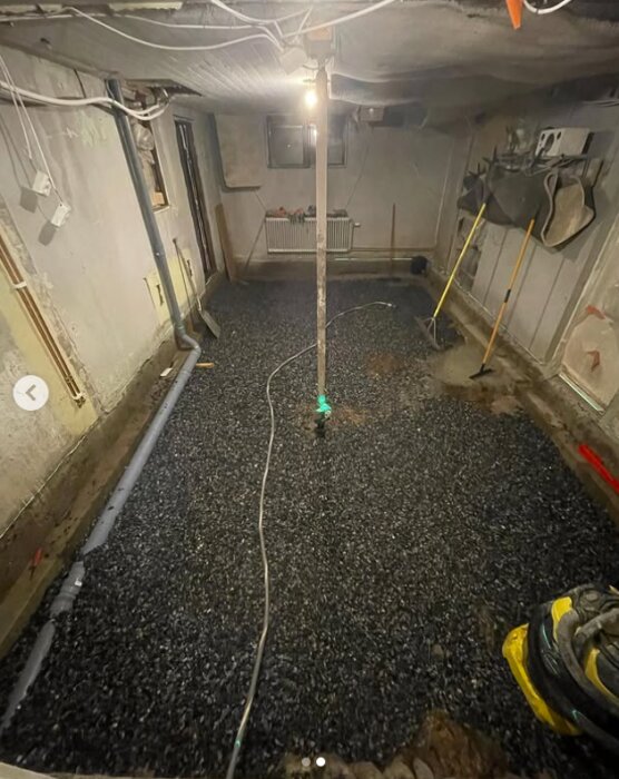 Källare med nylagd makadam, synliga rör längs väggen, verktyg lutade mot väggen och en avslagen bygglampa i mitten av rummet.