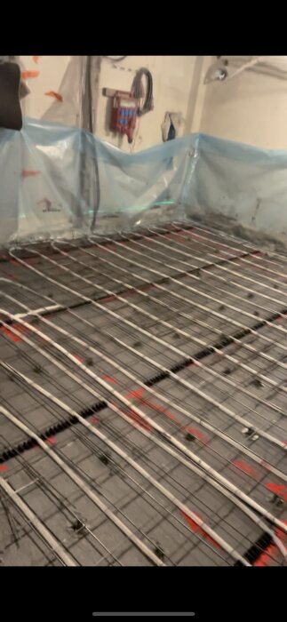 Byggarbetsplats med nylagd golvvärmeinstallation och skyddsplast på väggarna.