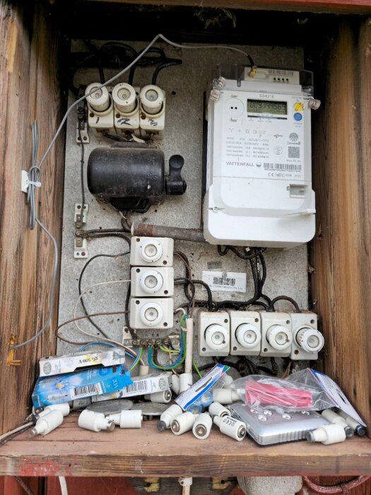 Elskåp utomhus med flera säkringar, en elmätare från Vattenfall och lösa säkringar samt förpackningar inuti, allt omgiven av en trästruktur.