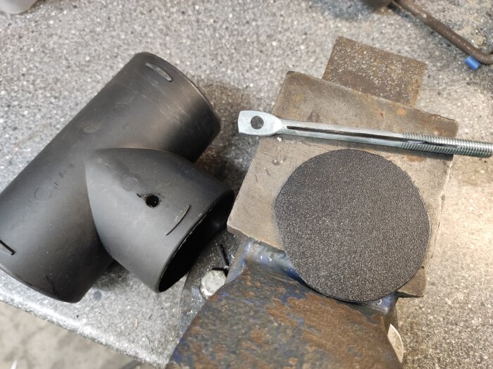 Svart Y-rör koppling och metallverktyg på bänk, bredvid en rund bit svart material.