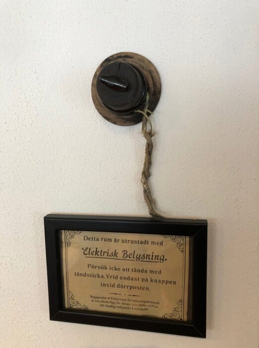 Gammaldags svart ljusknapp på väggen ovanför en inramad skylt som instruerar att vrida knappen i stället för att använda tändstickor för elektrisk belysning.