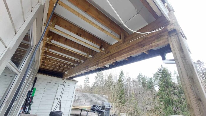 Utsikten under verandan visar träramar och bjälkar, med en stor mittdragare som sviktar något.områdets vegetation syns i bakgrunden.