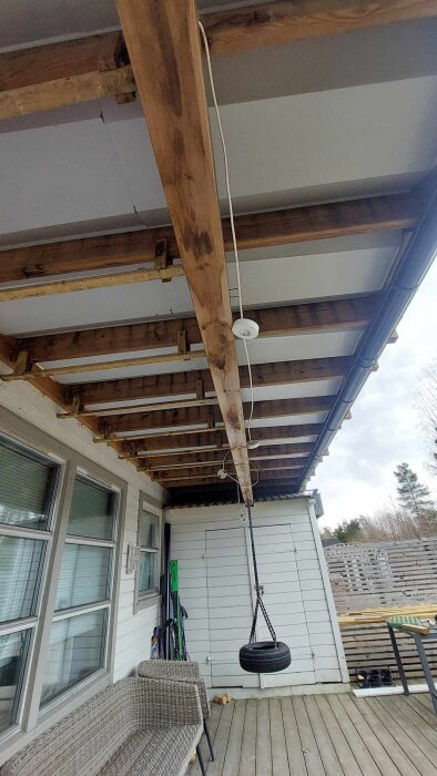 Utsikt under en veranda med träbjälkar, där en gunga hänger från taket. En sittmöbel och förvaringssaker syns i bakgrunden.