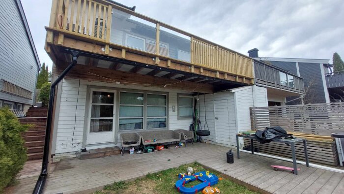 Träveranda med räcke ovanpå trädäck, sittgrupp, barnleksak och trädgårdsslang. Taket är utbyggt på verandan med böjd stödstruktur.