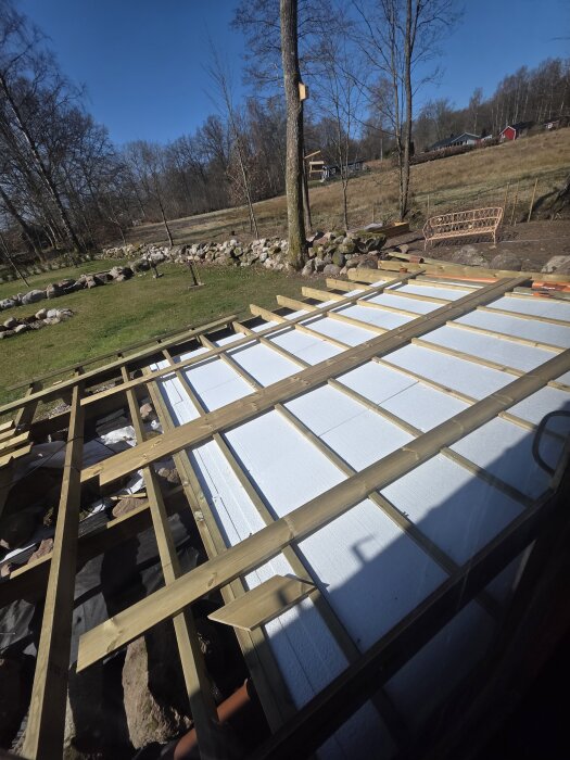 Träram under uppbyggnad för altan, med vit frigolitisolering placerad på marken, omgiven av träd och stenar i bakgrunden.