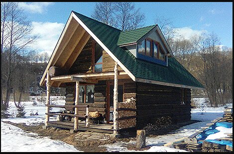 En liten timmerstuga med grönt tak och veranda står i ett snöigt landskap med några träd i bakgrunden.