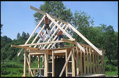 Bygge av hus med trästomme; tre personer arbetar med att montera takstolar på ett skelett av träplankor, omgiven av grönska.