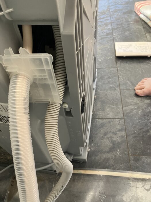 Baksida av vitvaror, inklusive en fäst avloppsslang och en delvis borttagen transparent panel, sett på ett klinkergolv.