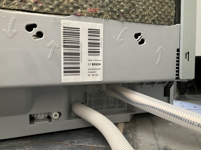 Grå panel på diskmaskin med fabriksvarningsetikett och pilar. Avloppsslangar är anslutna undertill.