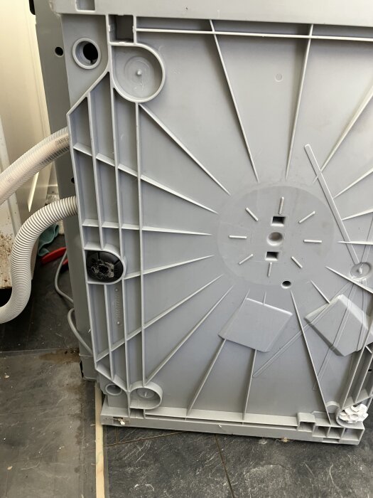 Grå panel på undersidan av en diskmaskin med avloppsslang synlig till vänster, visar svårigheter att ta bort panelen.