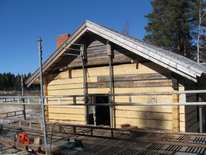 Byggnad med träbjälkar och tak under renovering, byggställning och verktyg synliga.