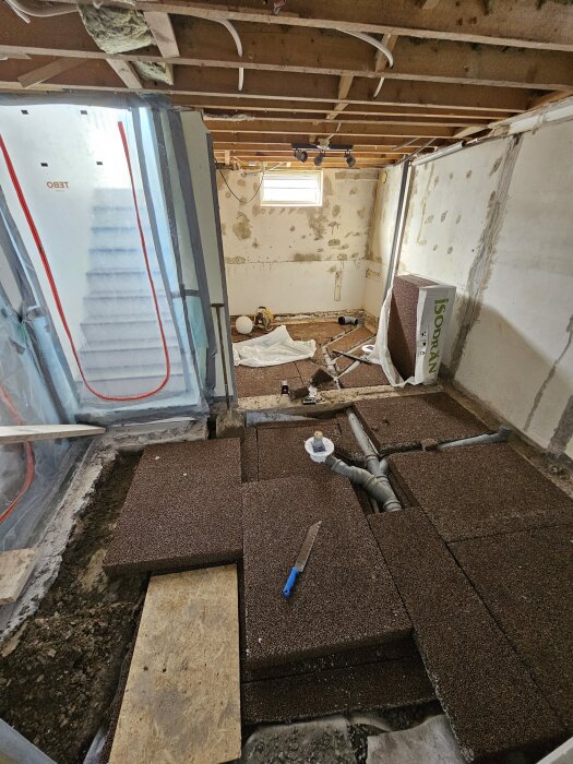 Isoleringsprojekt i källare med frigolitplattor på golvet, plaströr och en såg med blått handtag.