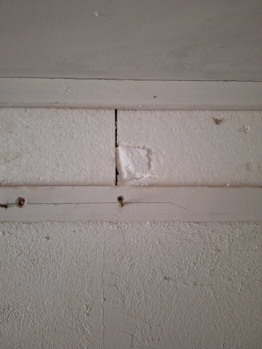 En frigolitskiva är monterad längs överkanten av en vägg med synliga skruvar och sprickor i ytan.