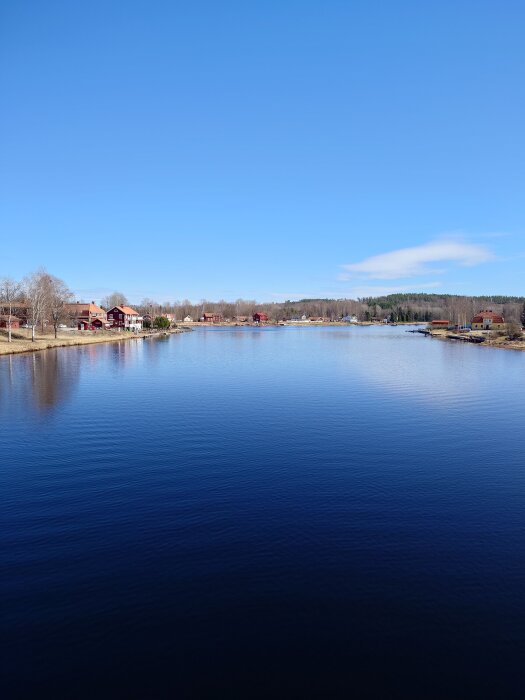 Sjön med klarblå himmel, omgiven av träd och traditionella röda hus längs stranden.