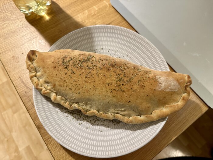 Inbakad pizza på en tallrik, dekorerad med örter, på ett träbord bredvid en dryck.