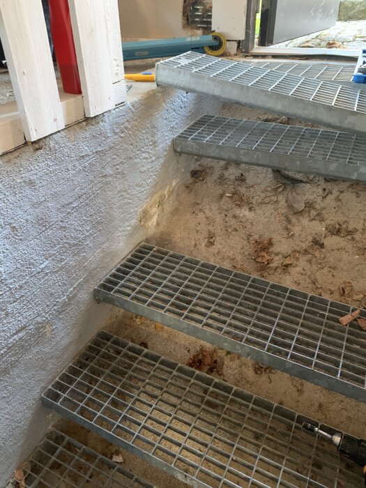 Lösa metallsteg i en utomhustrappa med en sluttande betongvägg syns i bilden, samt verktyg på marken i bakgrunden.