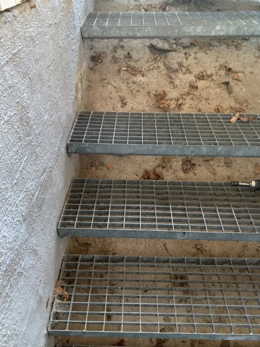 Metalltrappsteg på en källartrappa utomhus, delvis fasta, ovanför en betongslänt med viss jord och löv. Vägg i bakgrunden.