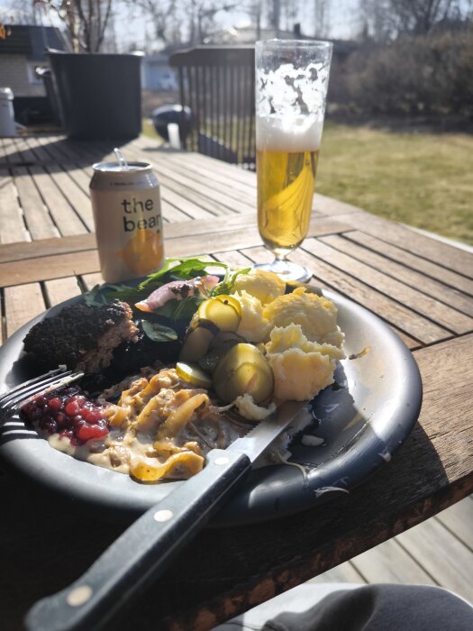 Tallrik med pannbiffar, hemlagat mos, gräddstuvad lök, lingonsylt, smörgåsgurka och sallad på bord, med öl och "the bear" burk.
