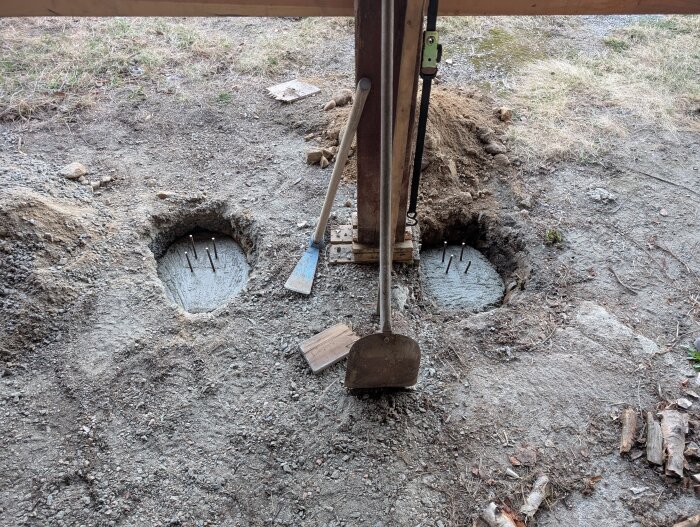 Jordhål med betong och järnpinnar, trästolpe som saknar markkontakt. Spade och skyffel bredvid. Förberedelse för väderskydd till traktor.