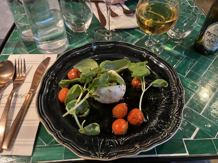 Svart tallrik med burrata, tomater och basilika. Grönt kakelbord, bestick och glas.