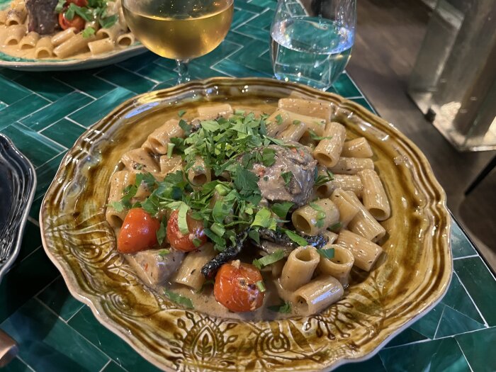Tallerken med Amici Caprece Rigatoni, toppad med körsbärstomater och örter, serverad med ett glas vitt vin på en grön bordsyta.
