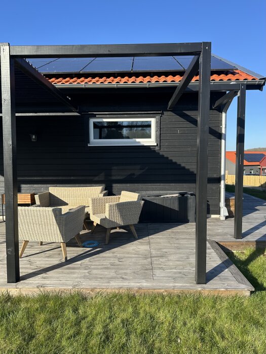 Uteplats med grå trallplattor, rottingmöbler och pergolastomme i trä. Svartmålad husfasad med solpaneler syns i bakgrunden.