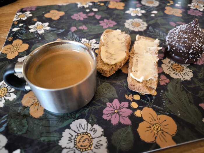 Kopp espresso, två grahamsskorpor med smör, och en chokladdragerad topp med kokosflingor på en blommig duk.