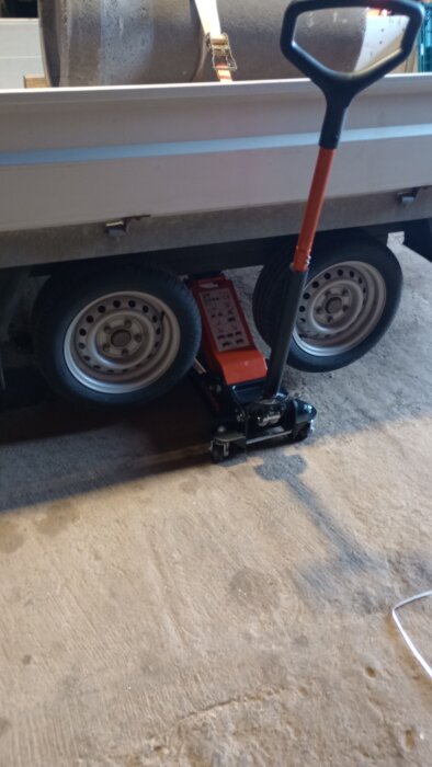 En orange domkraft under ett släp, redo att lyfta. Bakgrunden visar hjul och betonggolv i ett garage.