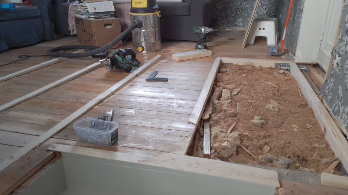 Golvrenovering pågår med uppbrutna plankor, isolering synlig, verktyg och skruvar i bakgrunden. Nytt golv förbereds.