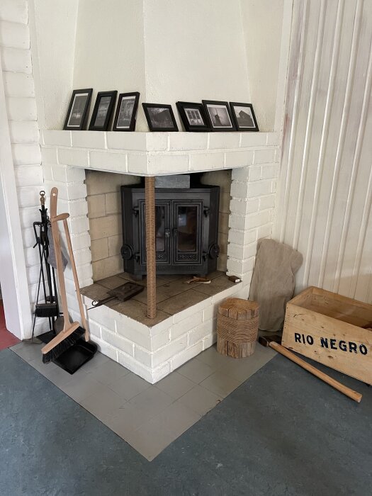 Vit murad hörneldstad med svart kamin, vedhink och verktyg. Fotografier ovanpå kaminen.