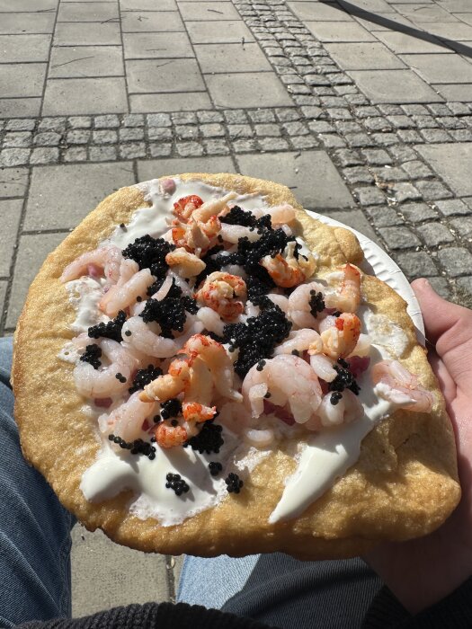 Langos med räkor, kräftstjärtar och svart kaviar, serverad på en tallrik, hålls i handen över en stenlagd yta.