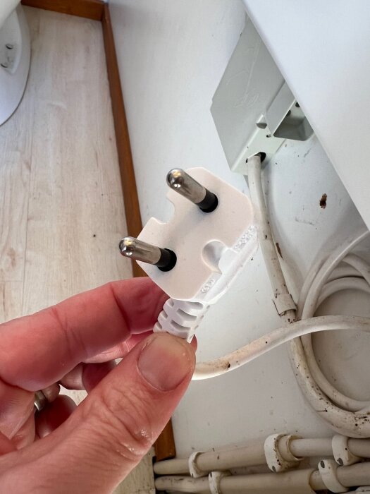 En hand som håller i en vit stickpropp med två stift framför ett eluttag vid en vägg.