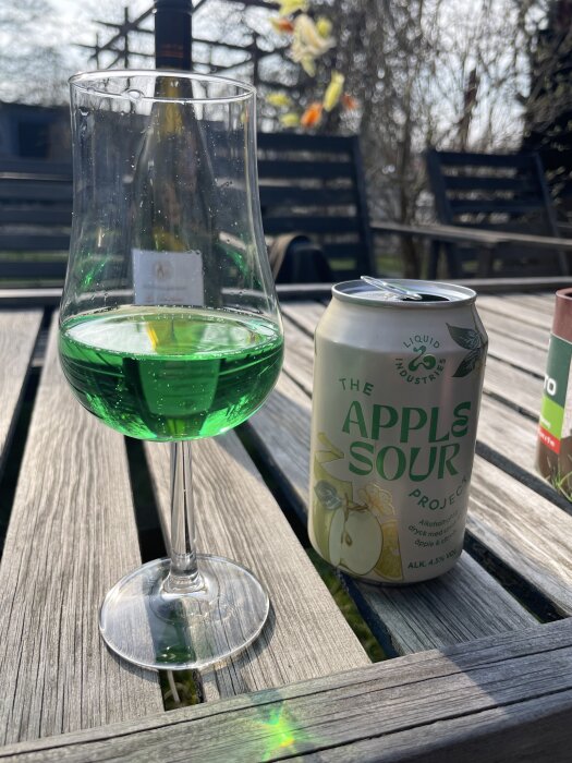 Grönt drinkglas och Apple Sour-burk på ett träbord utomhus i solen.