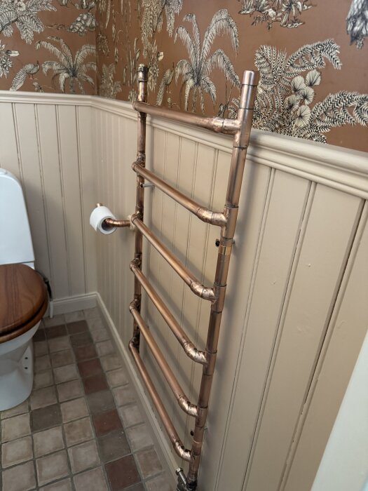 Uppvärmd handdukstork i koppar med flera lödda skarvar, monterad i ett badrum med mönstrad tapet och brunt kakelgolv.
