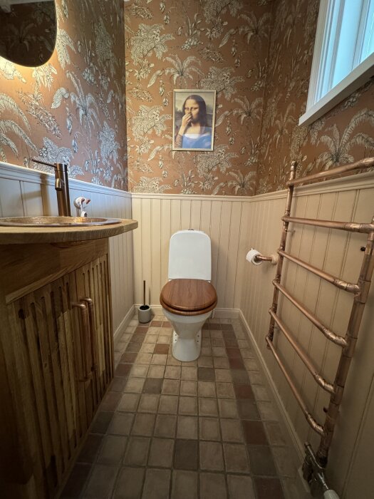 Badkar med tapetserade väggar, toalett, handfat i trä och kopparrörshanddukstork i ett badrum.