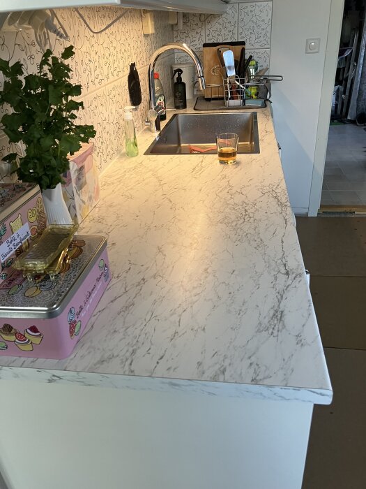 Kök med marmorbänk, diskho, kran, grönsaksväxt och olika köksartiklar på bänken. En drink står vid diskhon.