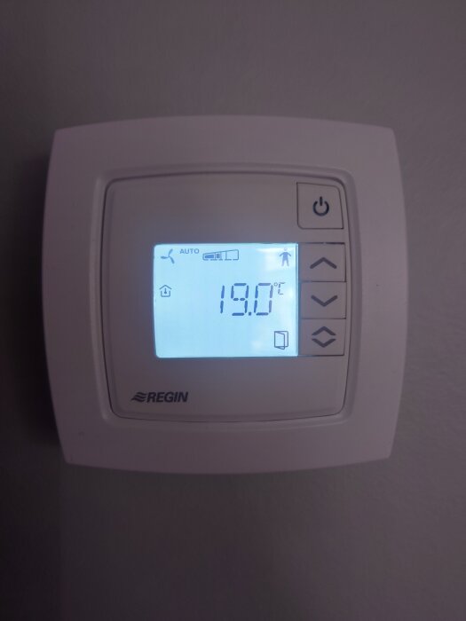 Digital termostat på väggen visar 19 grader Celsius, med symboler och knappar för inställningar.