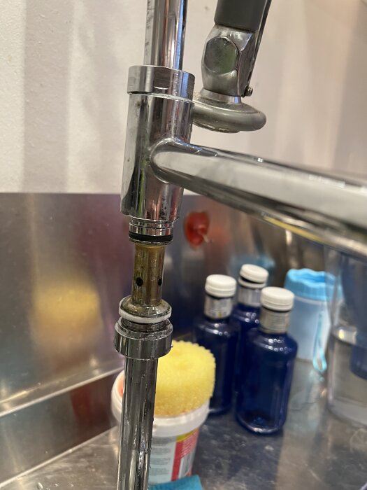 Närbild på en delvis demonterad köksblandare med synliga inre delar och en gummipackning. I bakgrunden syns flaskor och en rengöringssvamp.