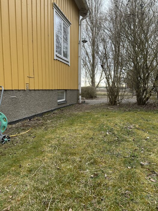 Gul husfasad med gräsmatta som lutar mot husgrunden. En trädgårdsslangvinda syns samt buskage i bakgrunden.