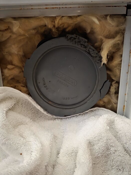 Ett trasigt täcklock av märket Wavin Access 110 omgivet av isolering och en handduk.