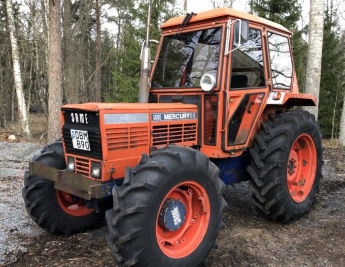 Orange SAME Mercury 85 traktor står parkerad i en skogsmiljö, med stora däck och hytt i metall.