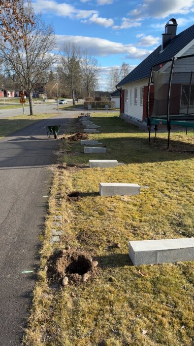 Grävd grop och staplade betongblock på gräsmatta nära hus, skottkärra i bakgrunden.