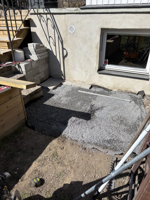 Utlagd stenflis på marken bredvid en husvägg med fönster, trappa och staplade murblock.