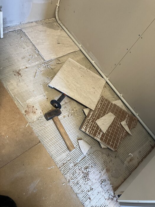 Krossade kakelplattor på golvet bredvid en hammare, redo för nästa steg i renoveringen.