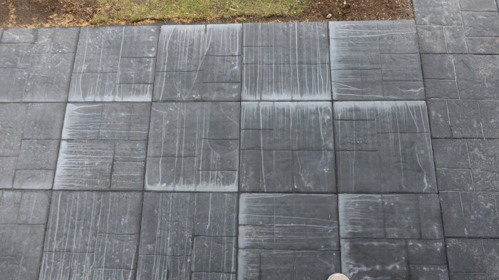 Gråa betongplattor med kalkutfällningar, synliga som vita fläckar, lagda på marken intill gräs.