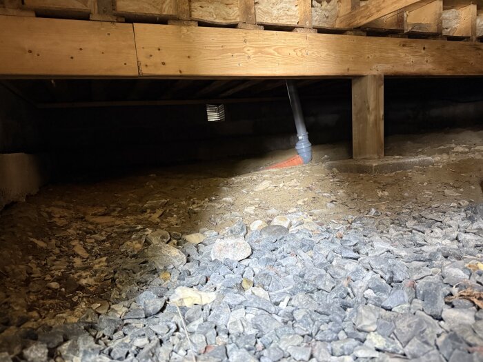Ventilation och plintar i krypgrund under utbyggnad, synliga träkonstruktioner och ett rör. Grus och jord på marken. Möjlig markfukt i fokus.